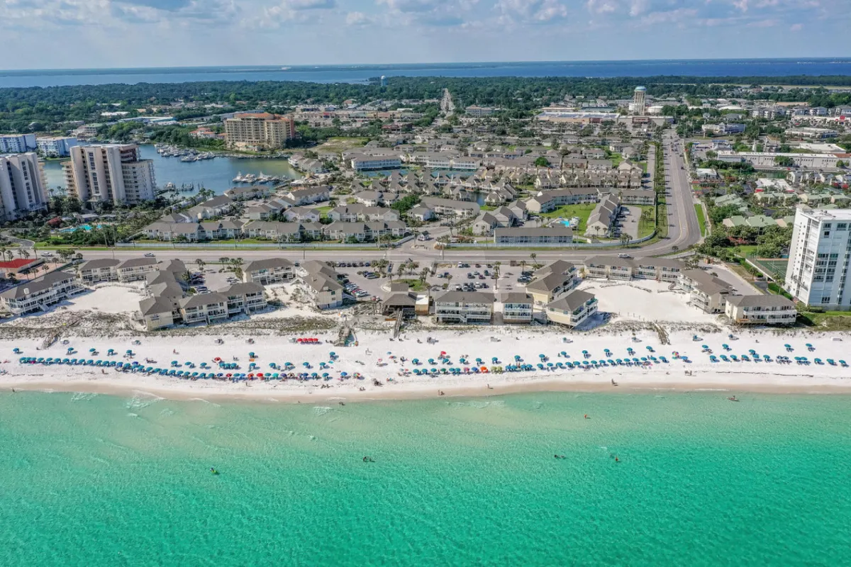 A beautiful stretch of pristine white sand beach bordered by clear turquoise water, with colorful umbrellas dotting the shore. Several buildings are visible in the background, indicating the close proximity of the accommodations to the beach. The scene feels relaxed and inviting, perfect for couples seeking a romantic morning stroll or a peaceful afternoon by the water.