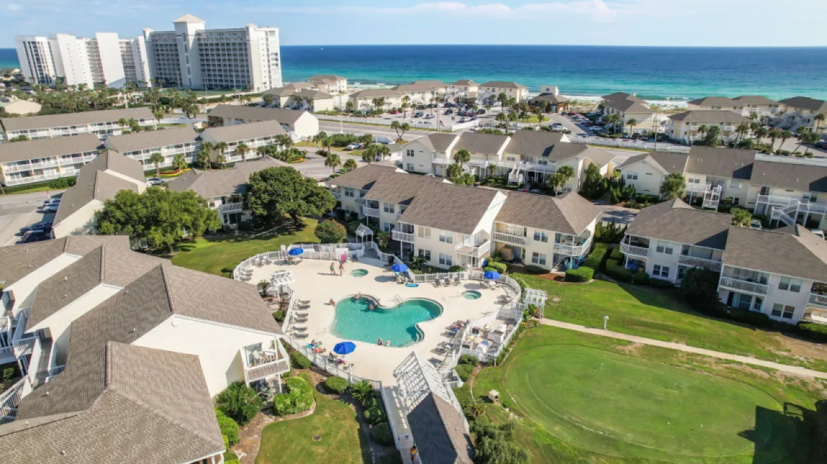 A lively resort scene featuring a sparkling swimming pool surrounded by lush green lawns, with sun loungers and guests relaxing by the water. In the background, the sandy beachfront and residential buildings capture the seamless connection between poolside leisure and immediate beach access.