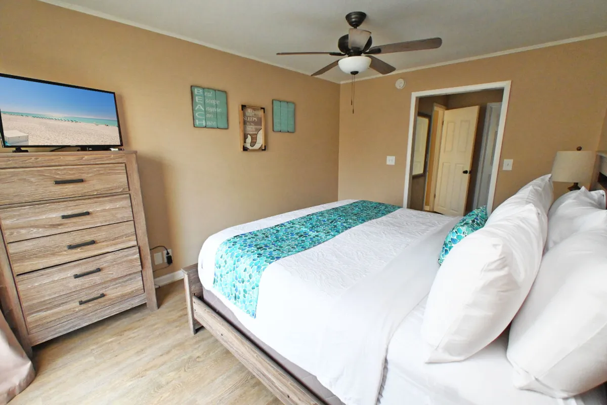 The image features a spacious bedroom with a bed adorned with turquoise accents, a wooden dresser, a smart TV displaying a tranquil beach scene, and decorative wall art. The soft lighting and carefully chosen decor create a calming, restful atmosphere ideal for unwinding after a day of activities.