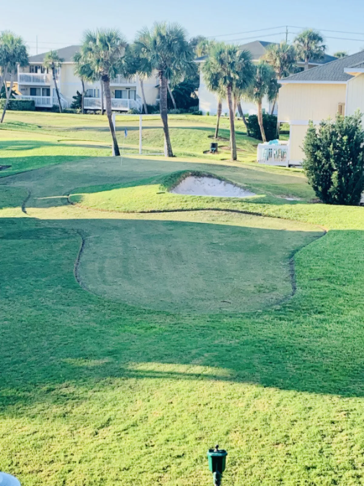 A scenic exterior view of the par-3 golf course, framed by palm trees and sand traps, with surrounding buildings in the distance. Golfers are seen enjoying a sunny day, capturing the spirit of recreation and camaraderie.