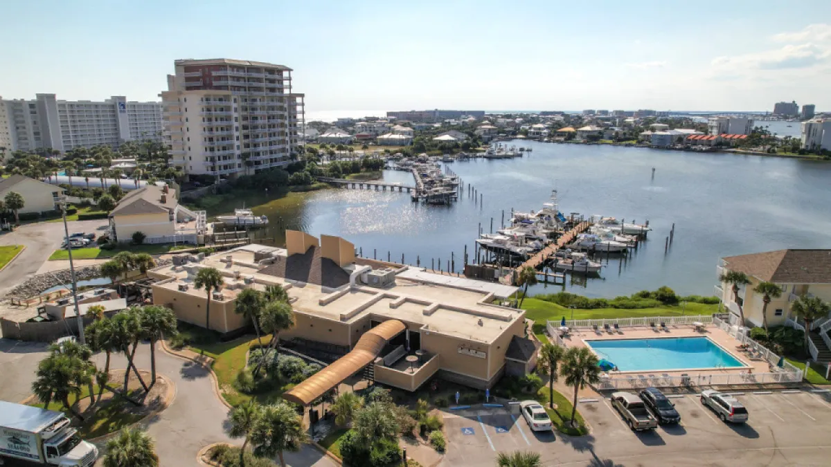 A sweeping view of the resort-style swimming pool sits in the foreground, surrounded by palm trees and manicured grounds. The water glistens in the sunlight, and a yacht dock is visible to one side, suggesting easy access to waterfront activities. The surrounding buildings hint at a close-knit, vibrant community.