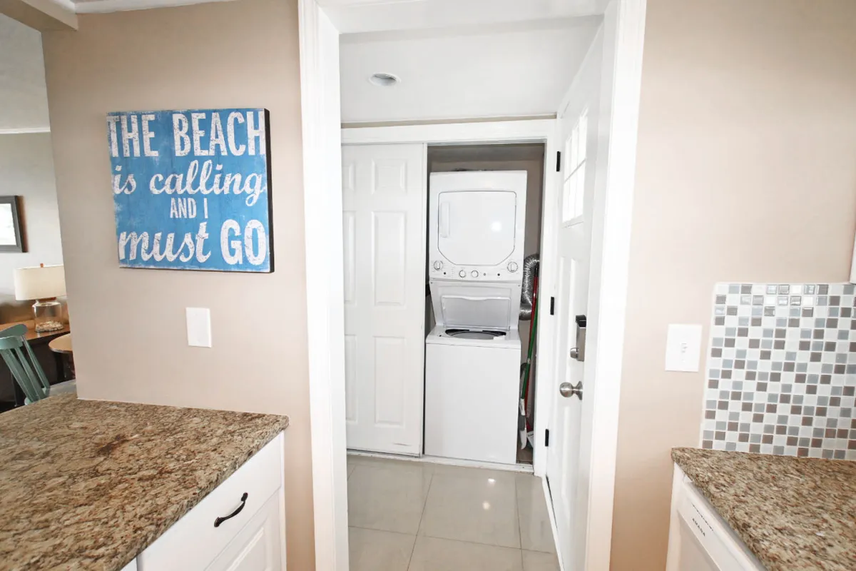 A cheerful corner featuring a laundry area with a granite countertop, a colorful backsplash, and a sign that reads 'The Beach is calling and I must go.' The washer/dryer is clearly visible, adding to the sense of home-like comfort.