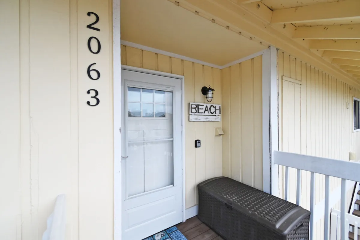 The image captures an entryway with a door labeled '2063,' a 'BEACH' sign, and a storage bench, evoking the sense of arriving home after a day at the beach. The bench hints at a practical space for stowing beach towels, chairs, and sporting gear.