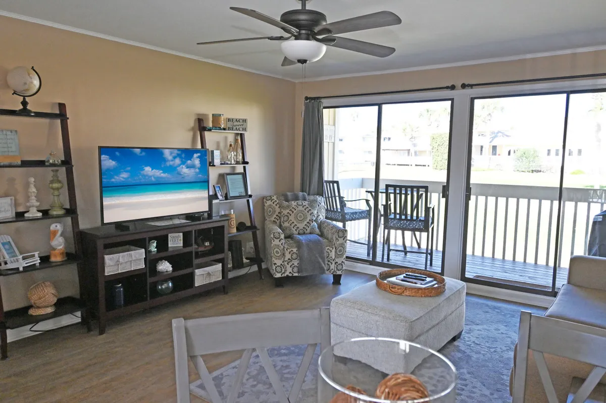 The image showcases a bright, inviting living room with a coastal-chic decor, featuring a plush sofa, patterned armchair, and a large flatscreen TV displaying a serene beach scene. A sliding glass door opens to a private balcony with outdoor seating, overlooking lush golf course views. Decorative shelving with beach-inspired accents completes the tranquil, resort-like ambiance.