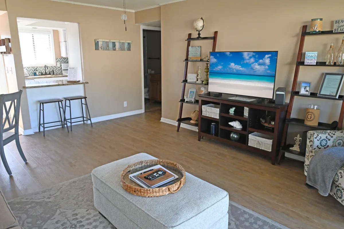 This interior image centers on a welcoming living area with a flatscreen TV displaying a beach scene, a kitchen bar with two barstools, decorative shelving with coastal accents, and a comfortable ottoman with a tray. The space feels open, bright, and cozy, ideal for relaxing after a day on the coast.