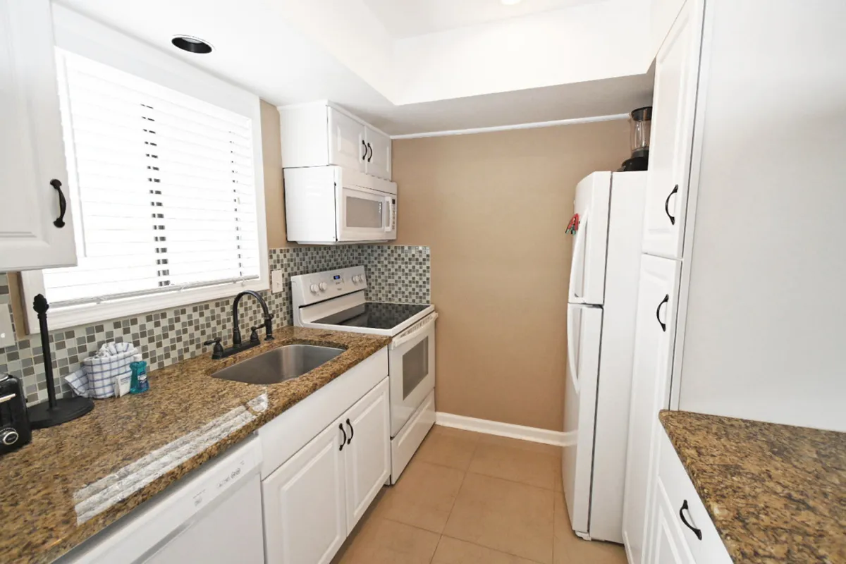 A bright, modern kitchen featuring white cabinetry, luxurious granite countertops, a modern stove, refrigerator, and a window letting in natural light. The space is organized and inviting, with appliances and ample counter space for meal prep.