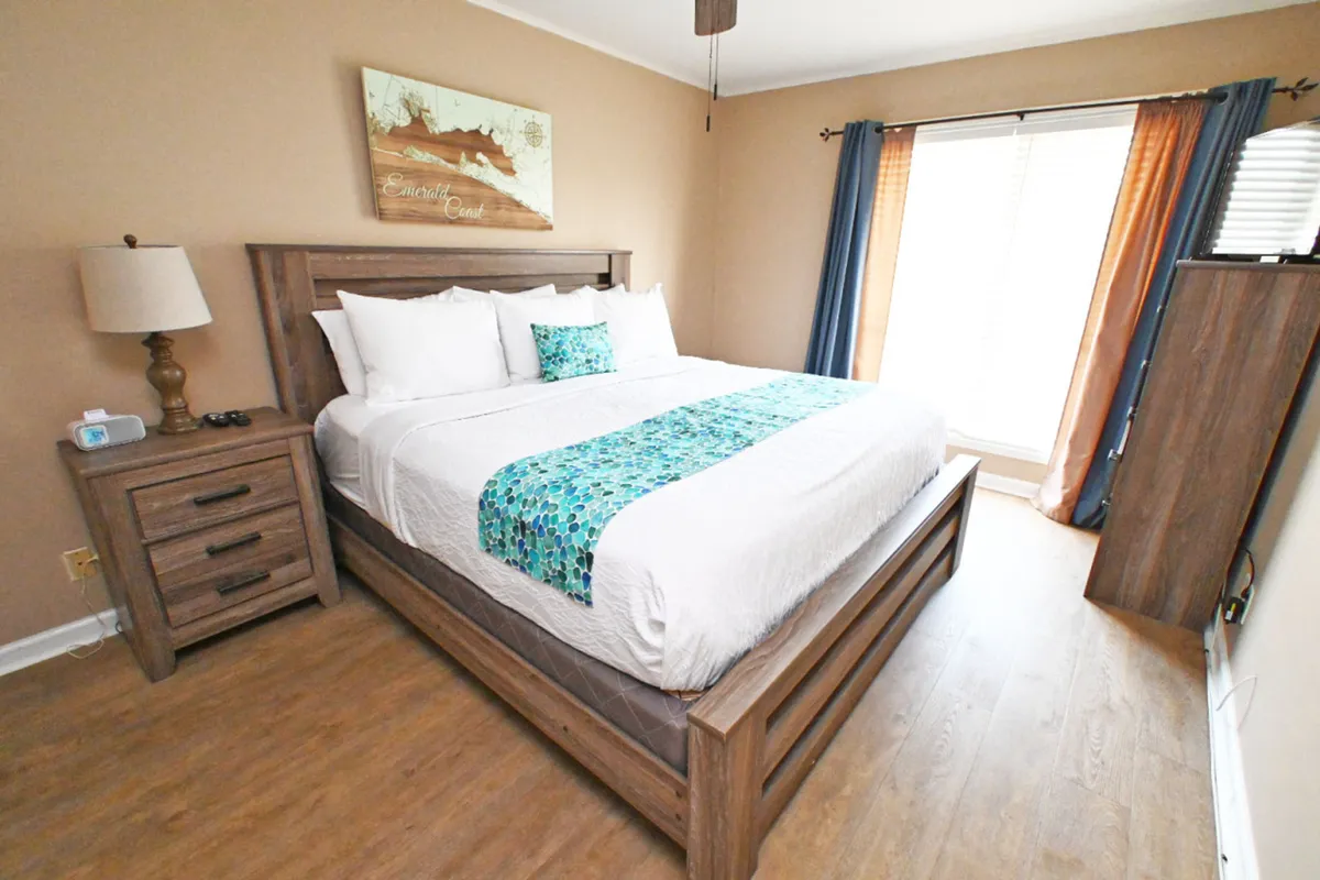 A spacious bedroom featuring a large king-size bed with crisp white linens and a turquoise accent runner. The room is brightened by natural light from the window adorned with blue and orange curtains. Beside the bed are a wooden nightstand with a lamp and a wooden dresser with a flat screen TV, creating an inviting and restful atmosphere.
