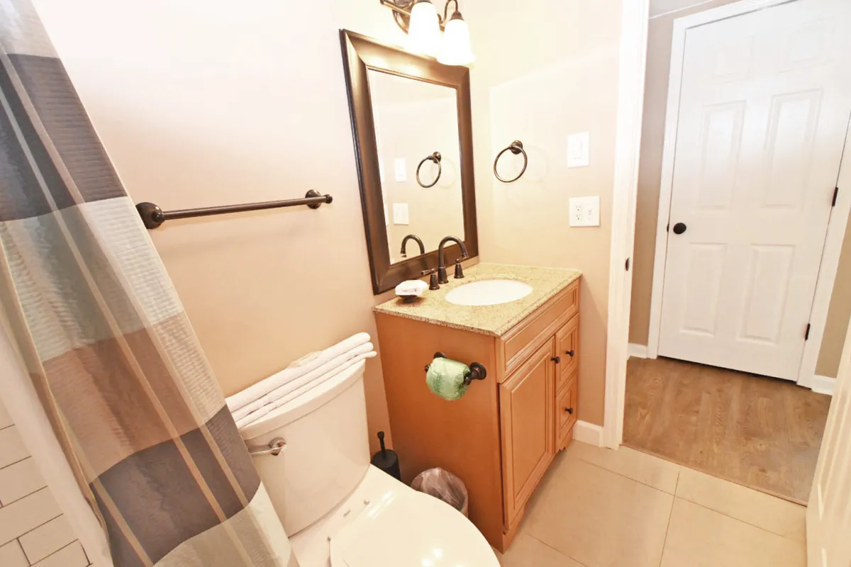A serene bathroom with a granite countertop vanity and sink, a large framed mirror, neatly folded towels on the toilet, and a shower with a striped curtain. The space is fresh, modern, and clearly updated for comfort and relaxation.