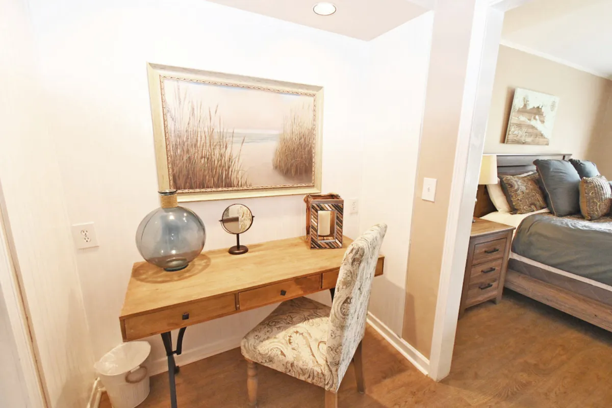 An inviting dressing area with a wooden desk adorned with decorative items and a patterned chair, complemented by coastal artwork. The view into the adjacent bedroom reveals a comfortable bed and bedside furniture, highlighting the functional transition between spaces.