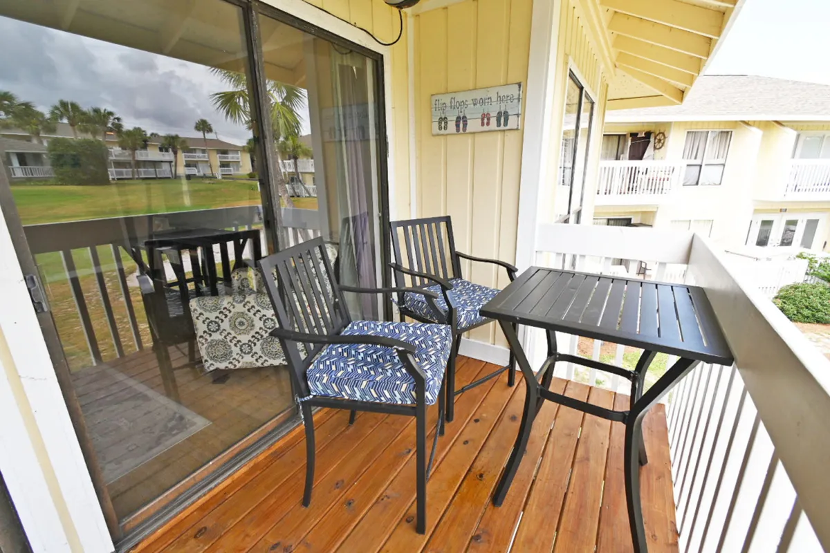 An exterior shot focused on a private balcony with a black metal table and two cushioned chairs, glass sliding doors reflecting the living room, and wooden floorboards. The setup suggests a tranquil spot for morning coffee or sunset cocktails while overlooking the greenery beyond.