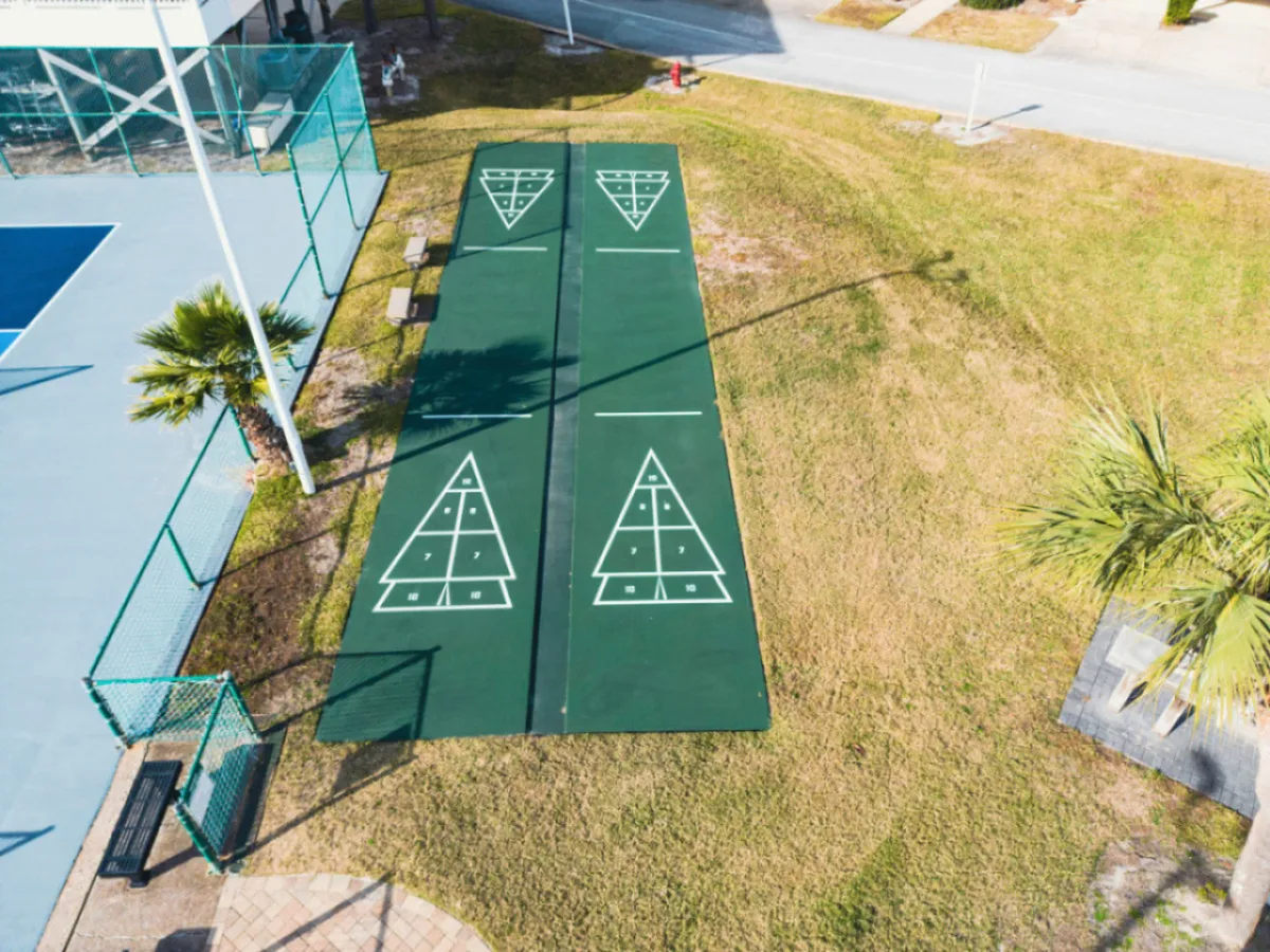 A set of shuffleboard courts with clear markings, a fenced tennis court with a bright blue surface, palm trees dotting the landscape, and benches for relaxing or socializing. The environment is cheerful and inviting, ideal for friendly competition and outdoor fun.