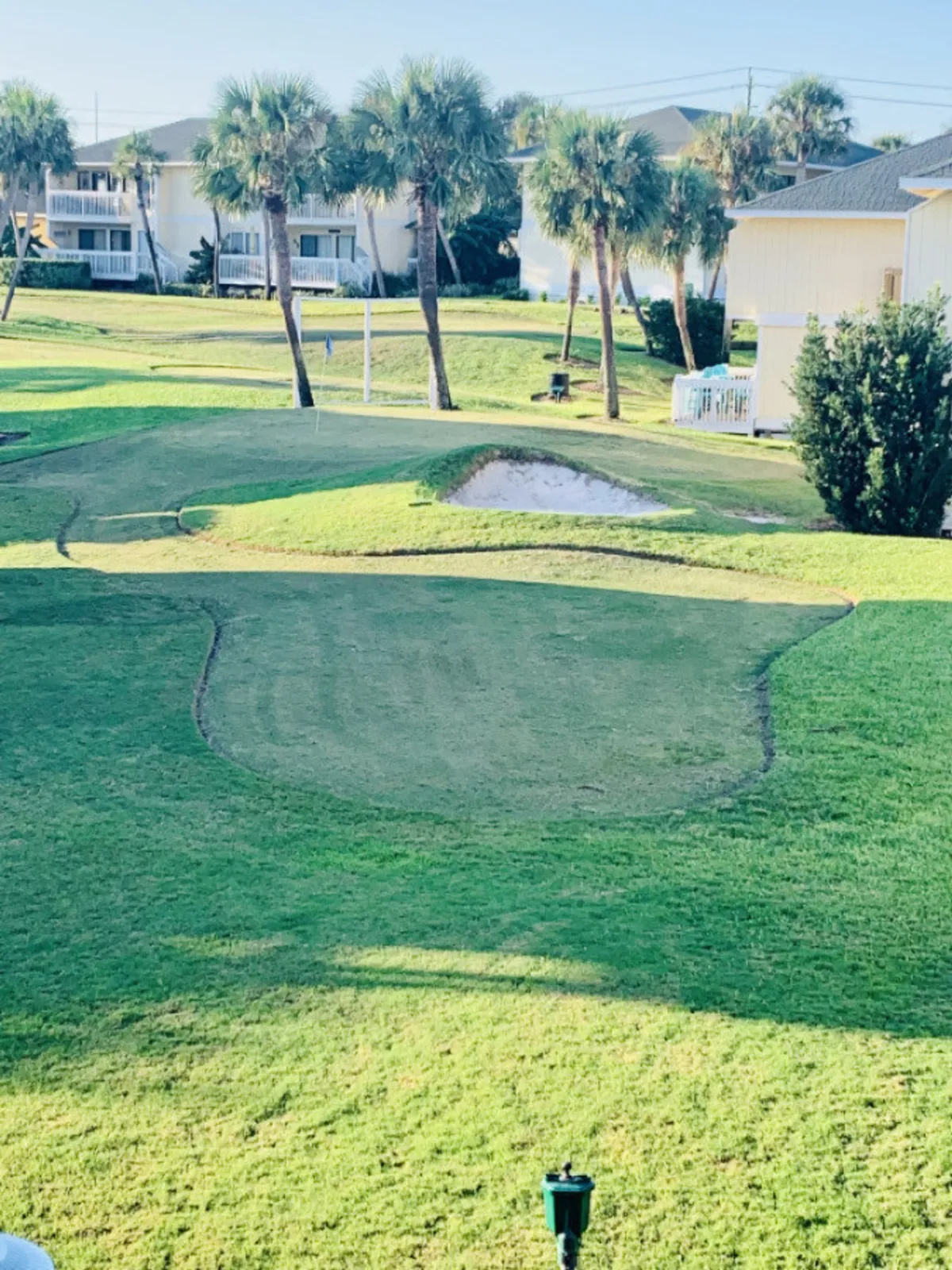 A lush, well-maintained par-3 golf course sits surrounded by tall palm trees and residential condos with balconies. The green is bordered by a sand bunker, and the setting is serene, inviting leisurely strolls or casual rounds of golf. The landscaping is vibrant, and the calm atmosphere is evident.