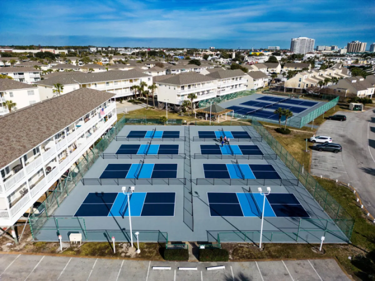 Multiple pickleball courts with vibrant blue and black surfaces, surrounded by condominium buildings and a parking area. People are actively playing on the courts, and others are socializing nearby. The scene is energetic and community-oriented, emphasizing active recreation.