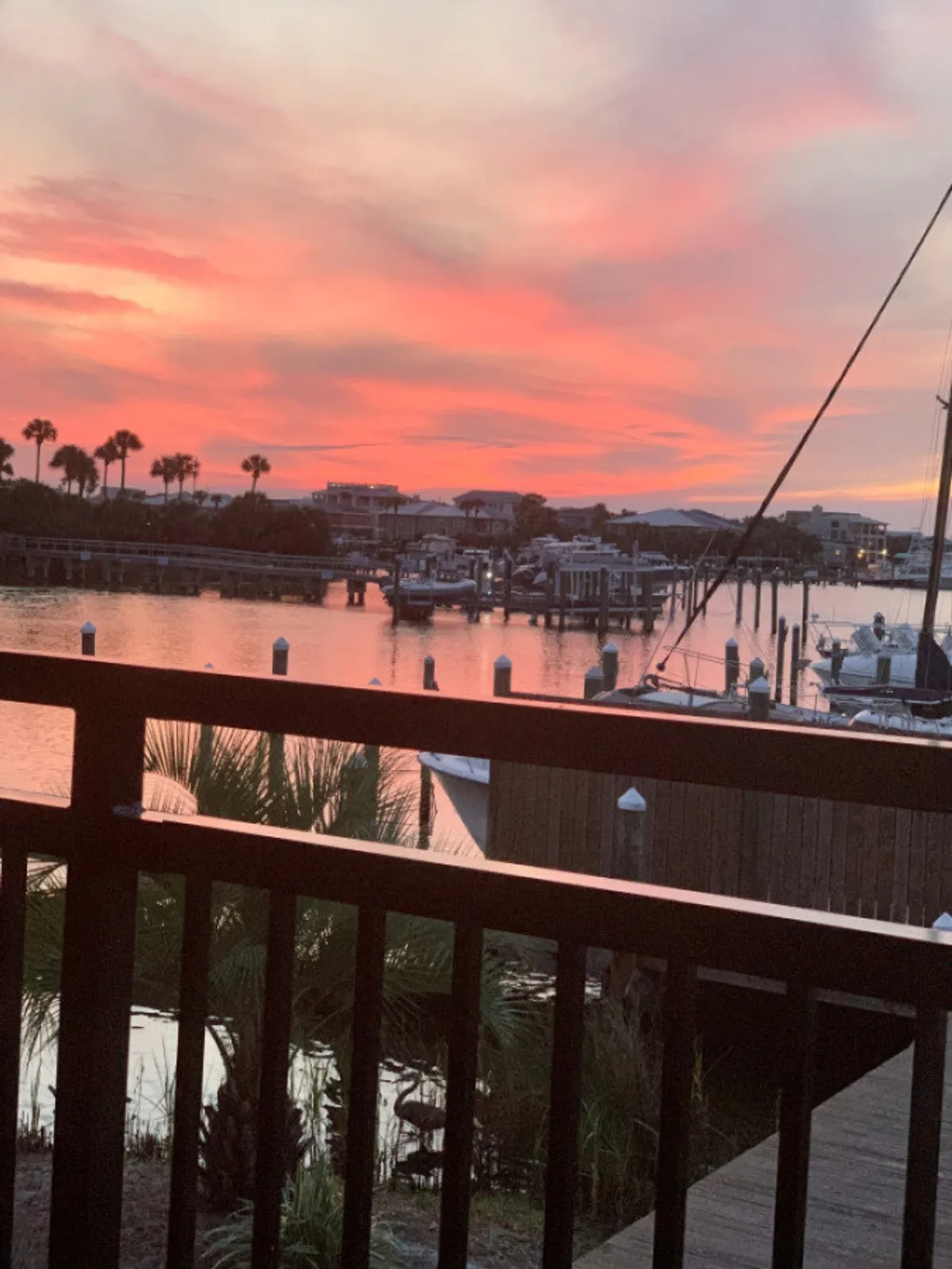 A vibrant sunset paints the sky above a marina filled with docked boats. The foreground features a wooden balcony railing, while silhouetted palm trees add a tropical touch. The peaceful water and tranquil atmosphere evoke the romance of evening waterfront moments.