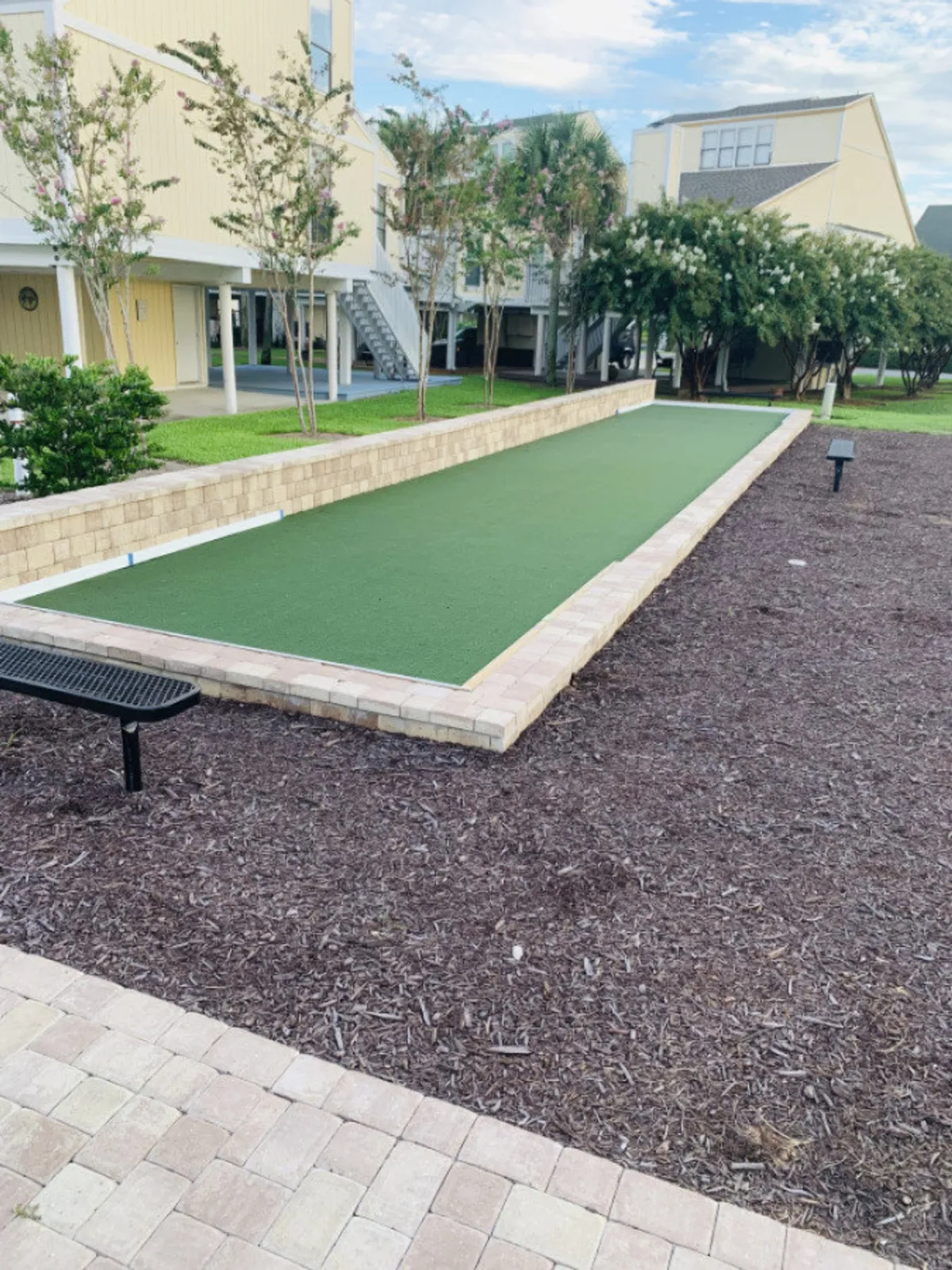 A bocce ball court with artificial turf is surrounded by manicured landscaping and residential buildings with balconies. A black metal bench sits nearby, offering a spot for relaxation or spectating.