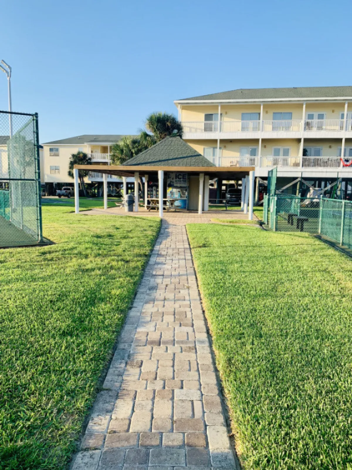 The scene features a brick pathway leading to a pavilion with picnic tables, flanked by fenced tennis courts on both sides. In the background is a multi-story residential building with balconies, all set in a calm, leisurely environment.