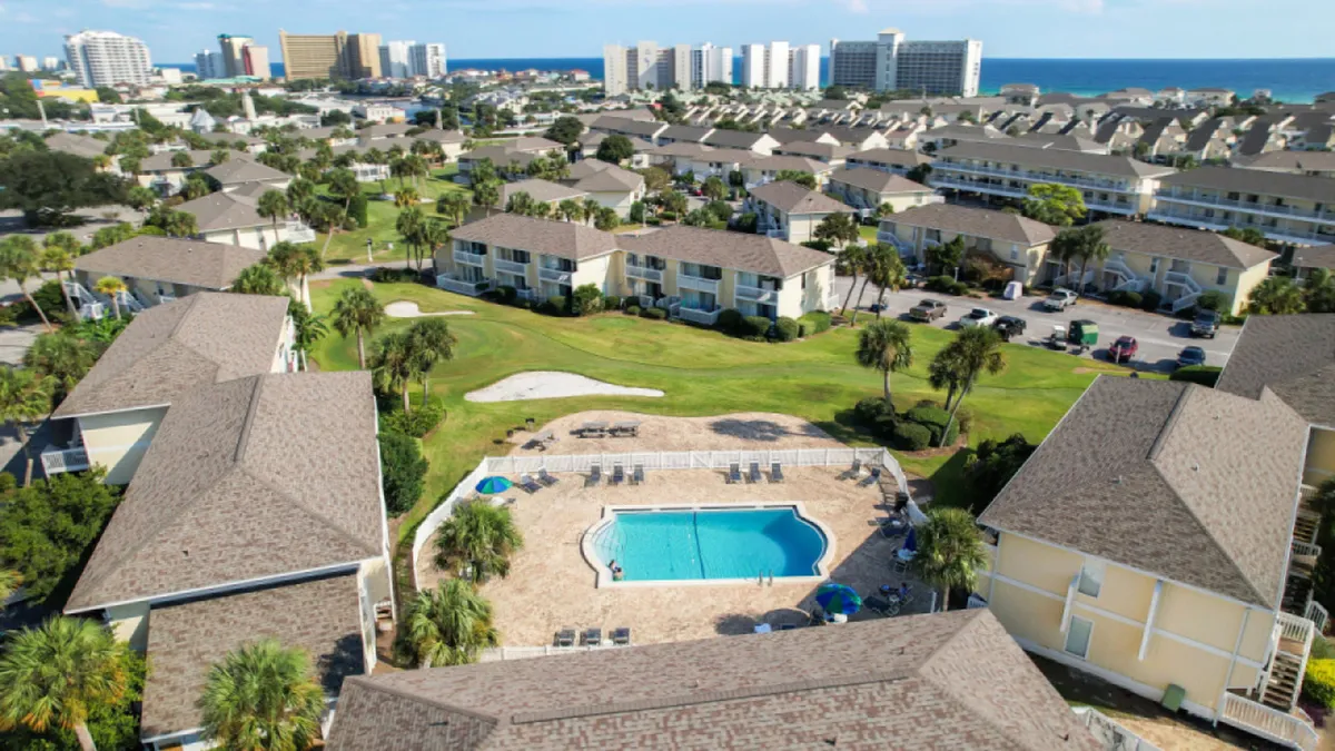 A central swimming pool surrounded by lounge chairs and condominium buildings with balconies. The pool area is bright and inviting, with green golf course fairways visible in the background and a distant view of high-rise buildings and the ocean horizon. The setting is perfect for family relaxation after a beach day.