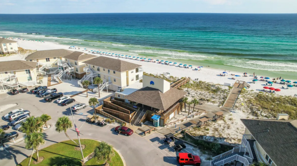 A pristine white sandy beach lined with colorful umbrellas and beach chairs, clear turquoise ocean water, a wooden boardwalk leading directly to the beach, and low-rise condos set back from the shoreline. The scene is lively with people sunbathing and enjoying water activities, reinforcing the beachside resort ambiance.