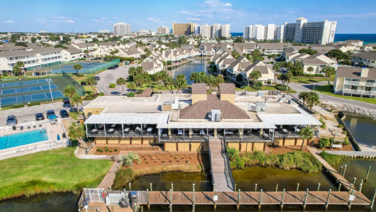 A vibrant exterior scene featuring the community clubhouse with covered patios, a sparkling swimming pool with lounge chairs, tennis courts, and a boat dock and marina area. The space is filled with opportunities for swimming, sunbathing, dining, and engaging in sports, all set against a backdrop of lush landscaping.