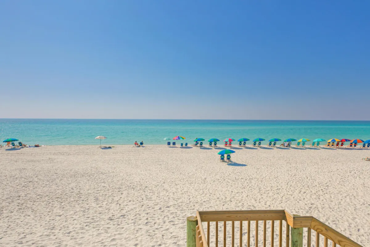 A wide, sugar-white sandy beach stretches into the distance, bordered by clear turquoise water and topped by a blue sky. Colorful umbrellas dot the shoreline, inviting visitors to relax. A wooden railing and stairs in the foreground hint at easy access from the community to the beach.