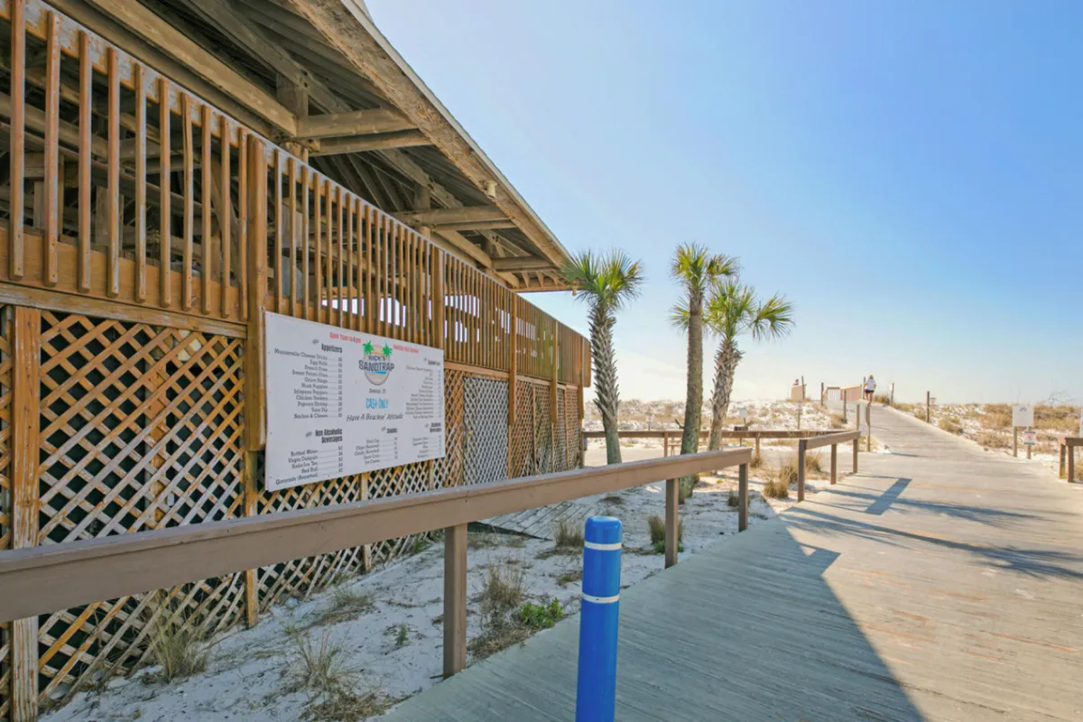 A wooden boardwalk leads through palm trees and dunes toward the private Destin beach. The boardwalk is flanked by sandy vegetation and a wooden structure with signage, capturing the calm and anticipation of a beach outing. The pathway feels tranquil and somewhat secluded, perfect for peaceful morning walks.